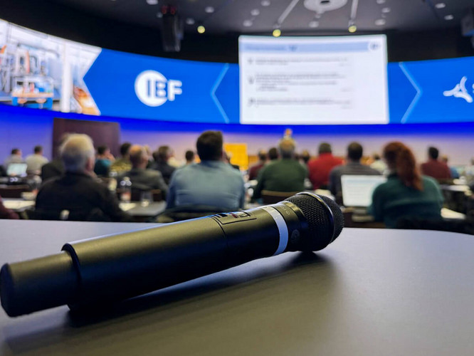 Ein Mikrofon liegt auf einem Tisch, im Hintergrund findet die Konferenz Maschinensicherheit 2025 in Zürich statt.