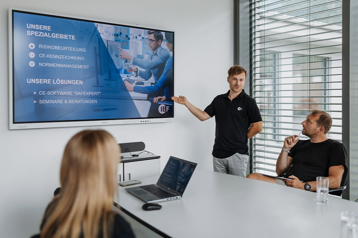Referent steht vor einer Gruppe Menschen und zeigt auf einen Monitor